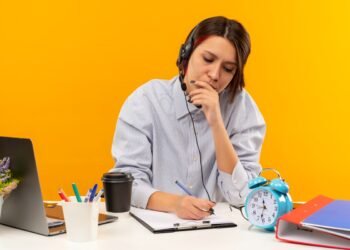 concentrated young call center girl wearing headset sitting desk putting hand mouth writing isolated orange 1 2