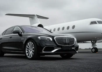 A glossy black luxury sedan positioned on the left side of the frame front angled towards viewer. To the right and background nose and fuselage of large white private jet visible resting on landing gear on 3