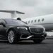 A glossy black luxury sedan positioned on the left side of the frame front angled towards viewer. To the right and background nose and fuselage of large white private jet visible resting on landing gear on 3