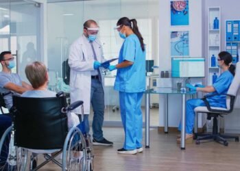 doctor with face mask against covid19 discussing with nurse hospital waiting area disabled senior woman wheelchair waiting examination assistant working reception computer 1